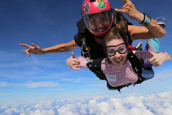 Female tandem skydiver wearing pink and white shirt giving thumbs up while in freefall.